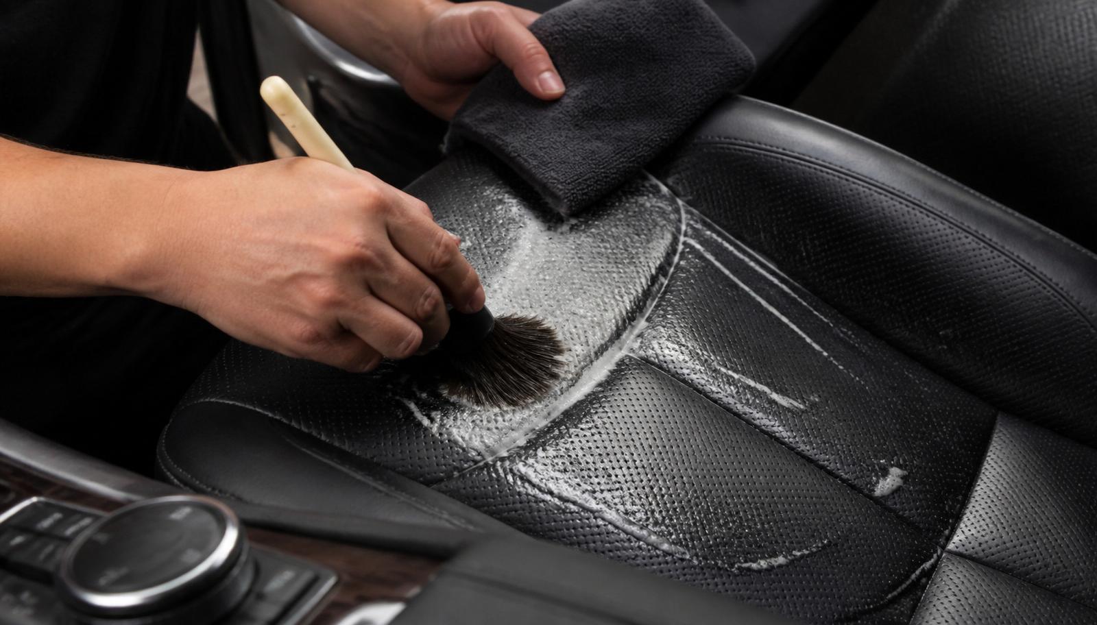Detailer cleaning a perforated black leather car seat with a soft brush and foam